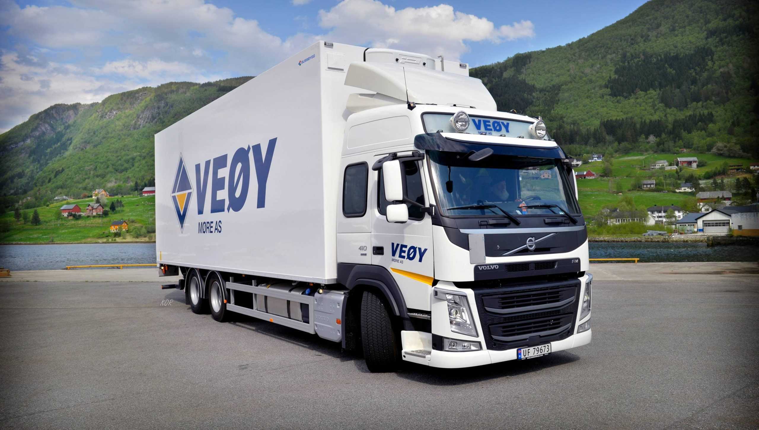 Stor hvit Volvo-lastebil med tilhenger merket VEØY Møre AS står parkert ved fjorden, omgitt av grønne fjell og små hus i bakgrunnen.