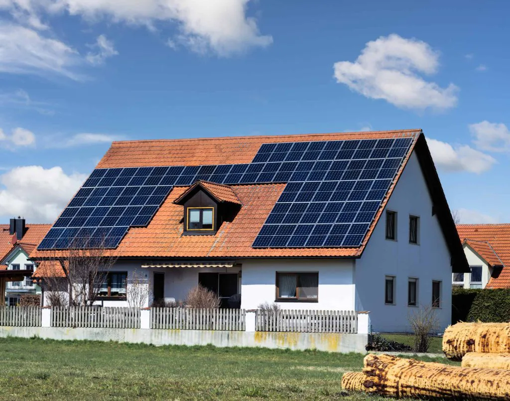 Enebolig med hvit fasade og stort oransje tak dekket av mange solcellepaneler; et lite loftsvindu i taket. Foran huset grønt gress og hvitt gjerde, med en stabel vedstabler i forgrunnen til høyre.