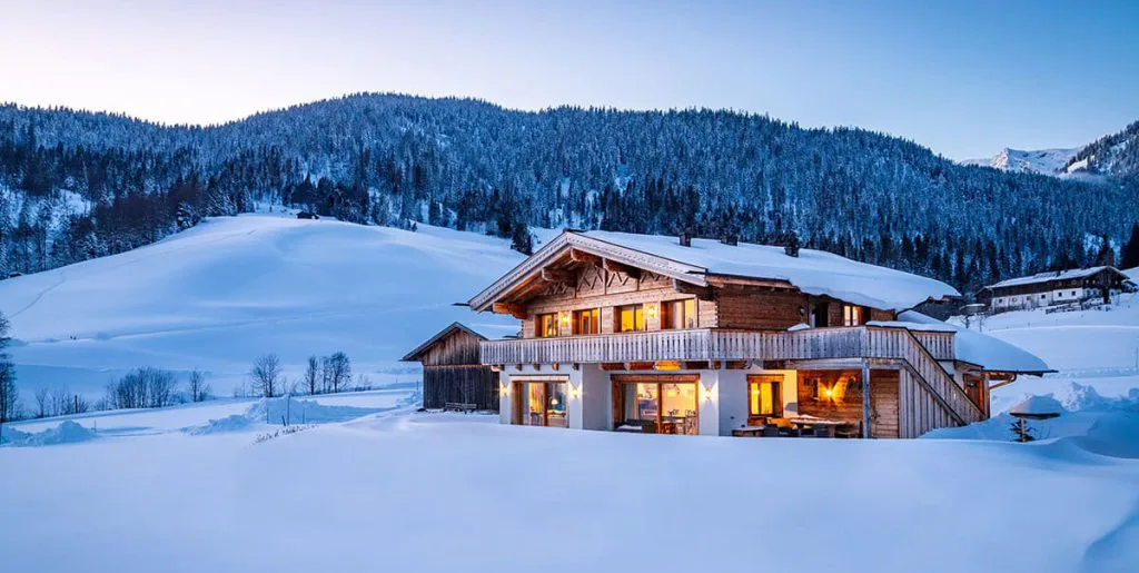 Koselig trehytte i snødekt landskap med varmt lys i vinduene og balkong, omringet av snødynger og skogkledde fjell i bakgrunnen.